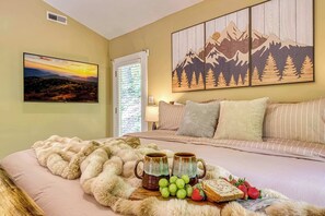 A bedroom with a bed, a painting of mountains, a window with blinds, and a tray with fruit and mugs. - Pine and Peak – Hot Tub, Fire Pit, Dog Friendly (Gatlinburg)