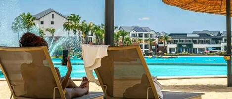 Two lounge chairs under thatched umbrellas by a pool with a view of resort buildings.
