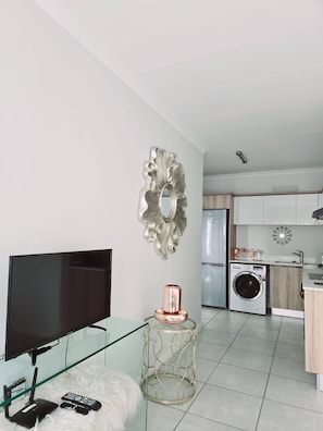 A modern living room with a flat-screen TV, a glass coffee table, a fluffy white rug, a decorative mirror, and a kitchen area with a washing machine.