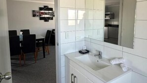 Un baño moderno con azulejos blancos, un lavamanos, un espejo y una puerta que da a un comedor.