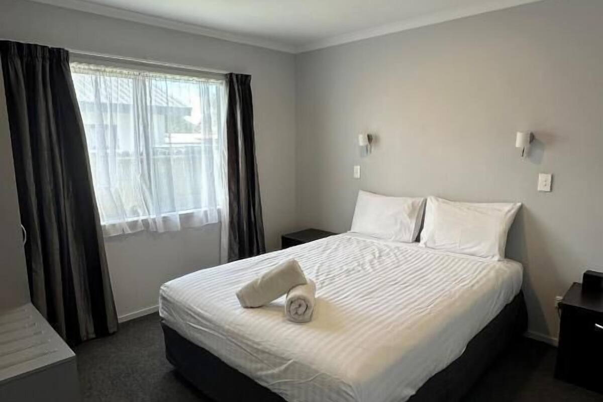 A hotel room with a bed, a window with curtains, a bedside table, and a wall-mounted light.
