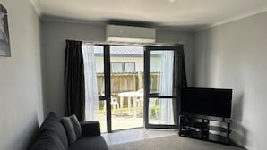 Una sala de estar con un sofá, un televisor de pantalla plana sobre un mueble bajo y un ventanal corredizo que da a un área exterior.