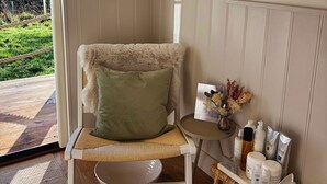 A chair with a fluffy pillow, a small table with skincare products, and a view of a wooden deck.