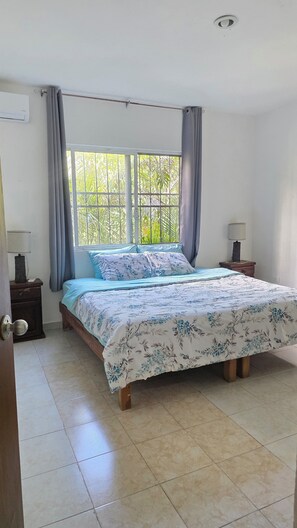 Un dormitorio con cama, cortinas, ventana, mesita de noche y lámpara.