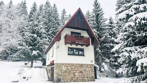 A snow-covered house with a stone foundation and a red roof, surrounded by snow-laden trees. - Chalet Helena by Interhome (Nové Hamry)