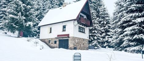 A snow-covered house with a stone foundation, a sloped roof, and a small balcony.