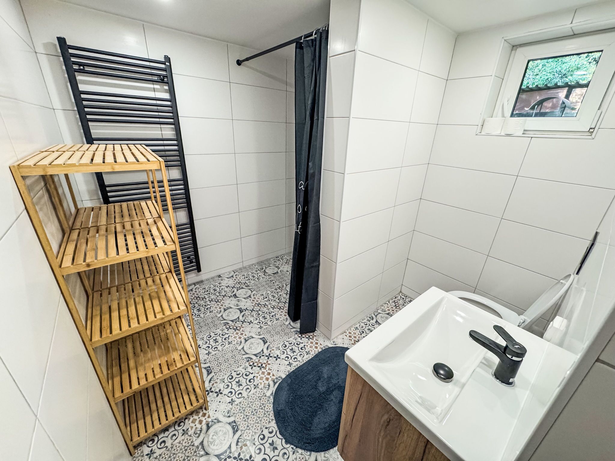 A bathroom with a shower, a wooden shelf unit, a sink, and a window with a view.