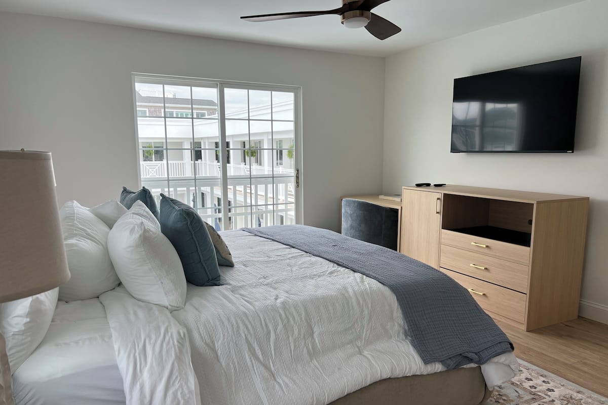 Un dormitorio con cama, televisor, escritorio y ventana con vistas a edificios.