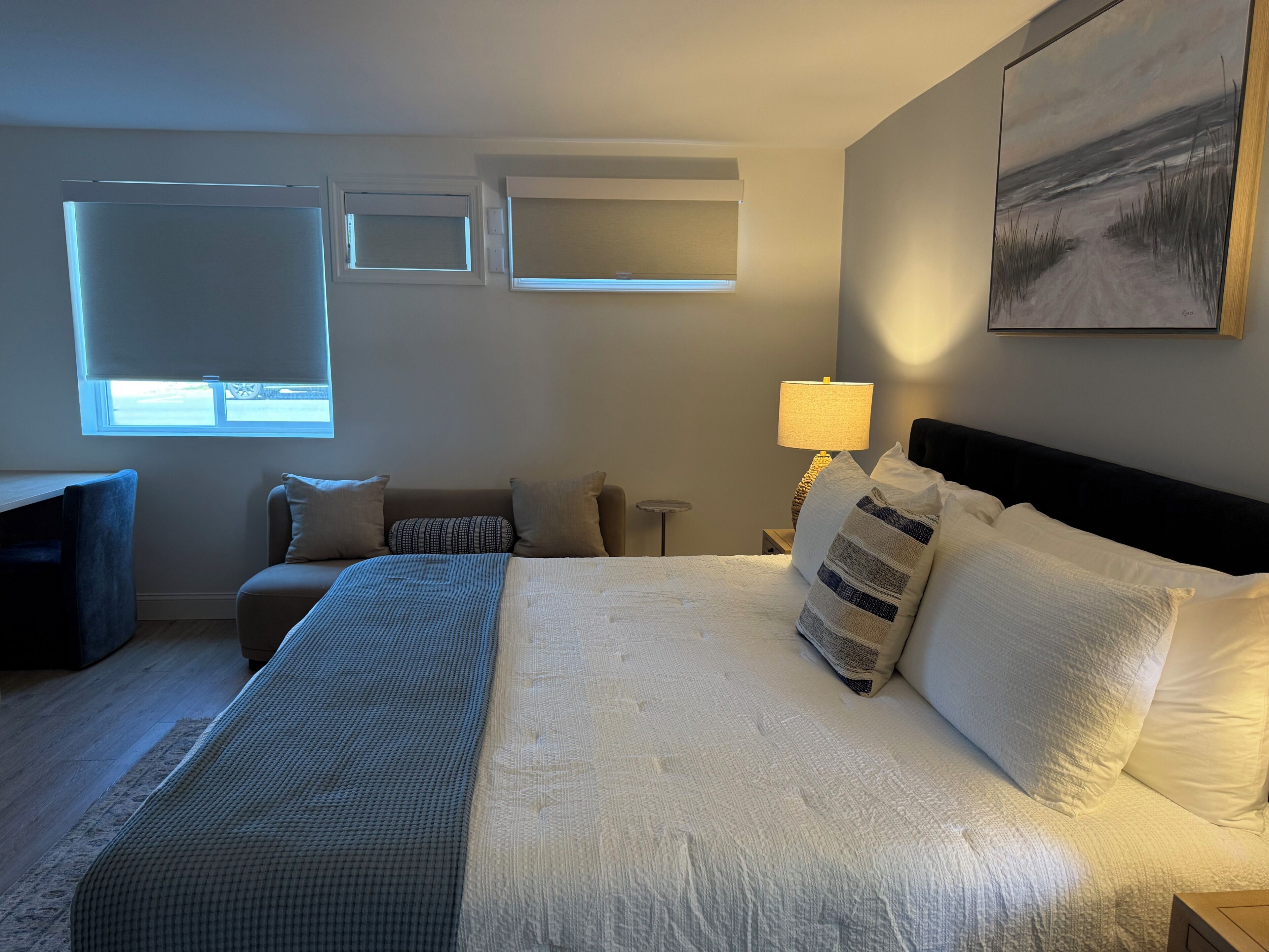A bedroom with a bed, a desk, a chair, a lamp, and a painting on the wall.