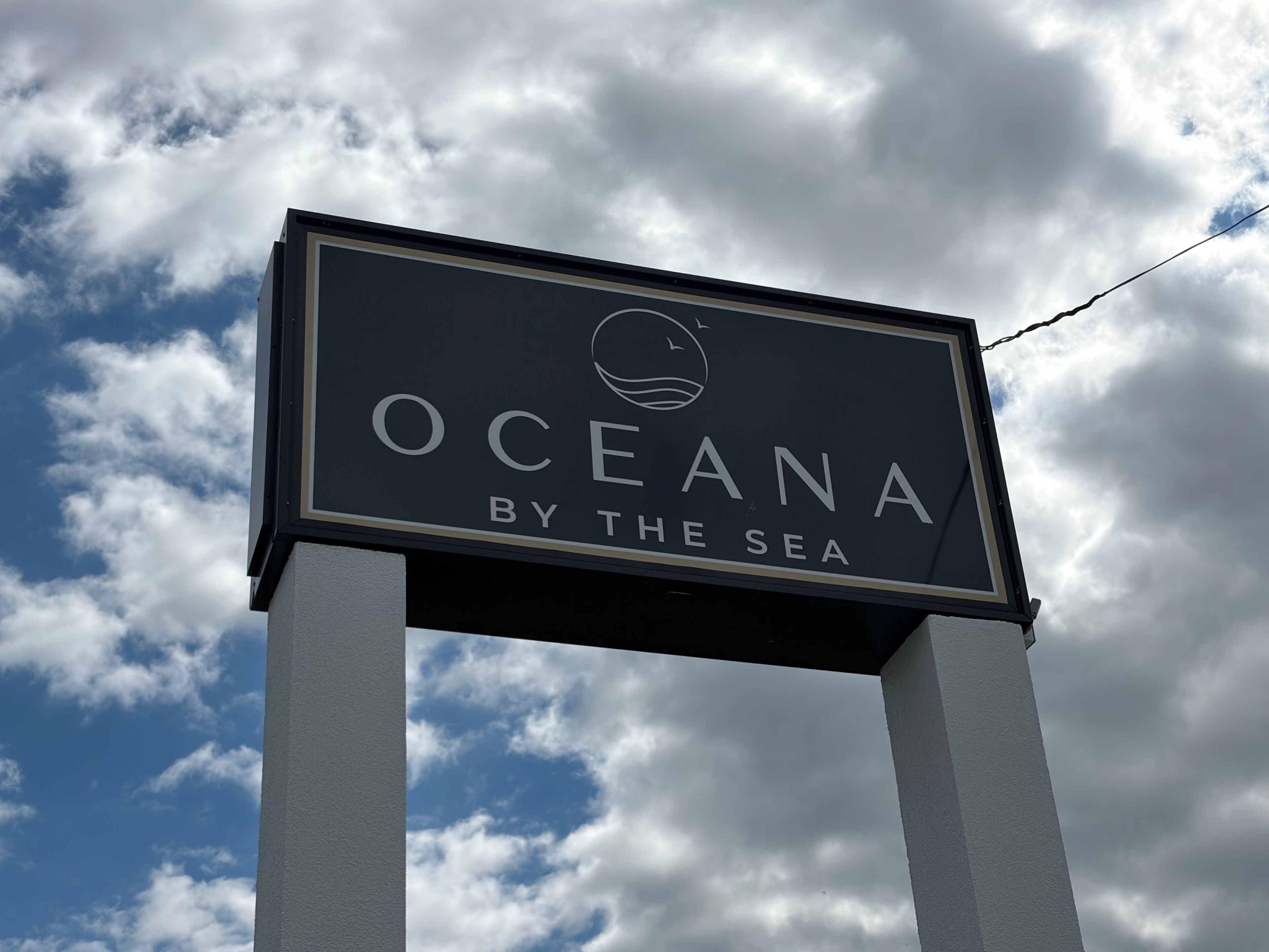 Un cartel con el texto 'OCEANA BY THE SEA' frente a un cielo parcialmente nublado.