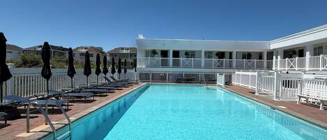 Una piscina rectangular con un indicador de profundidad de 1,8 metros, rodeada de sillas de descanso y sombrillas, con una valla blanca y un edificio al fondo.
