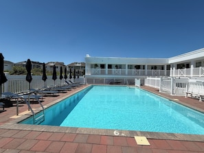 A rectangular swimming pool with a 6 ft depth marker, surrounded by lounge chairs and umbrellas, with a white fence and a building in the background. - Oceana By The Sea (Point Pleasant Beach)