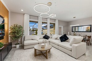 A modern living room with a sectional sofa, a round coffee table, and a large window with curtains. - Brand New | 5BR Pool Home w Safari Loft 16200 (Clermont)