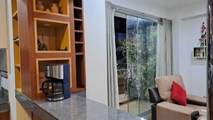 Una cocina moderna con cafetera, un mueble de madera con estantes y un ventanal corredero que da a un balcón con plantas.