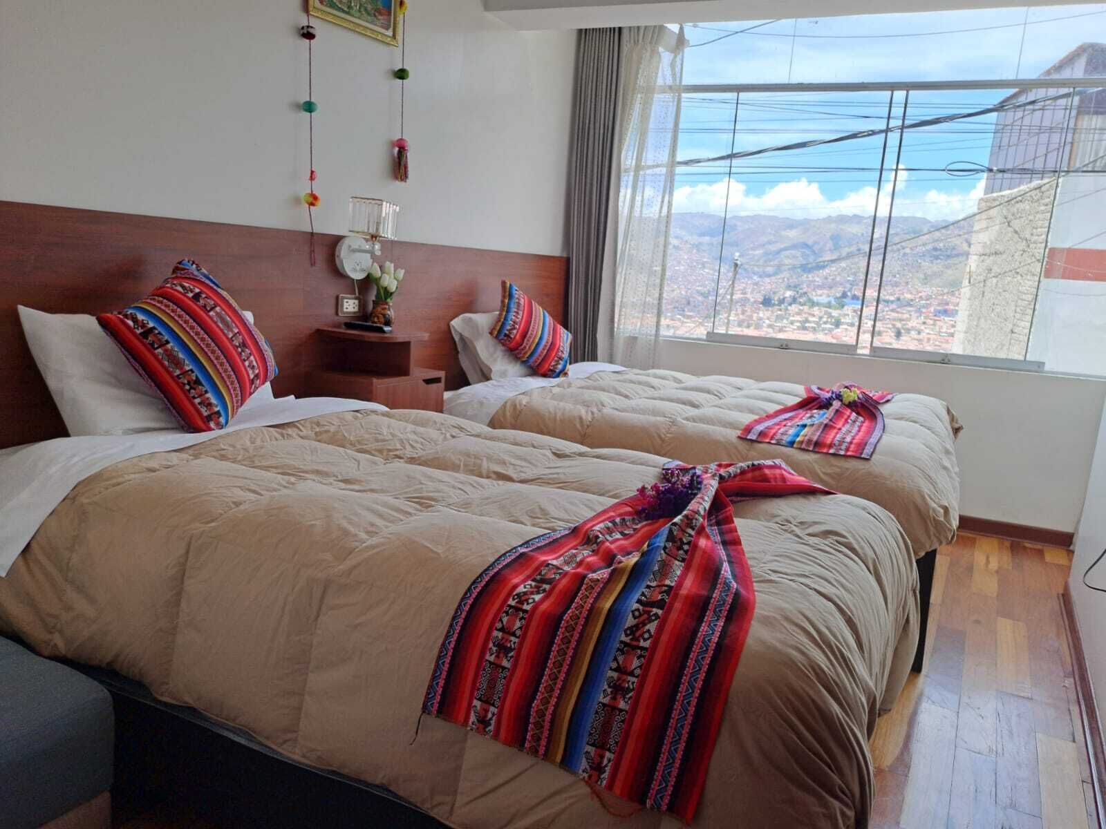 Habitación de hotel con dos camas, cabecero de madera, mesita de noche con un reloj, un florero con flores y un ventanal con vistas a la montaña.