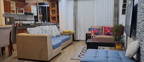 A modern living room with a sofa, a coffee table, and a TV. There is a kitchen area with a refrigerator and a window.