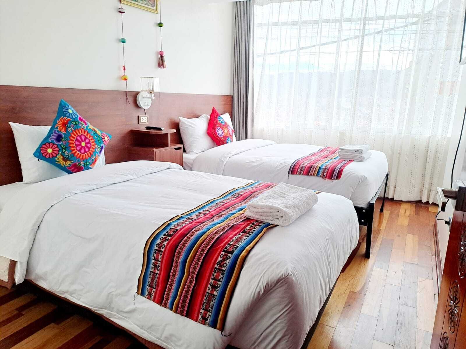 A hotel room with two beds, white bedding, colorful pillows, a bedside table with a lamp, and a window with sheer curtains.