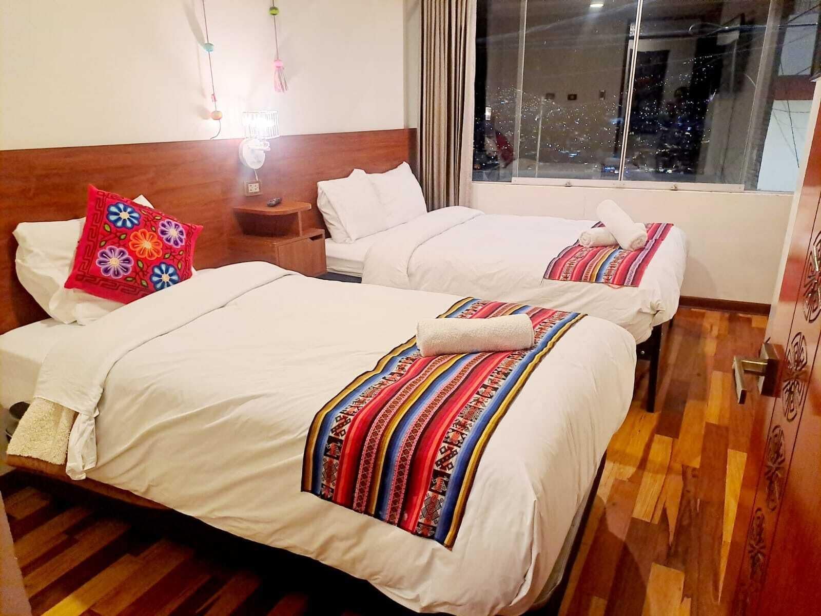 A hotel room with two beds, a nightstand, a window with a view, and a wall-mounted light fixture.