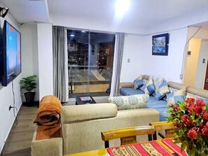A living room with a sofa, a table with a red tablecloth, a TV mounted on the wall, and a sliding glass door leading to a balcony. - RESIDENCIAL CRISTINA  (Cusco)