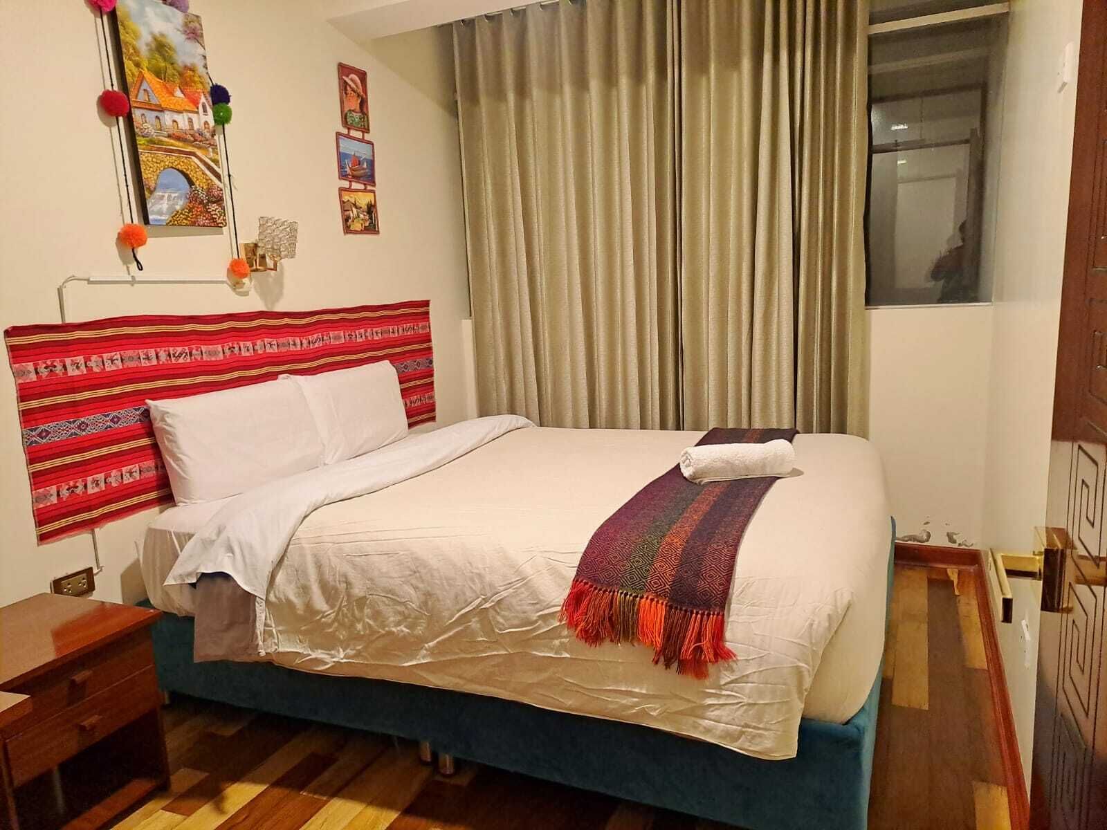 A neatly made bed with a white comforter and a colorful blanket, a wooden nightstand, a wall with framed pictures, and a window with curtains.