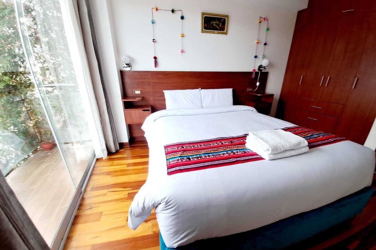 A hotel room with a wooden floor, a bed with white bedding, a wooden wardrobe, a nightstand, and a window with a view of trees.
