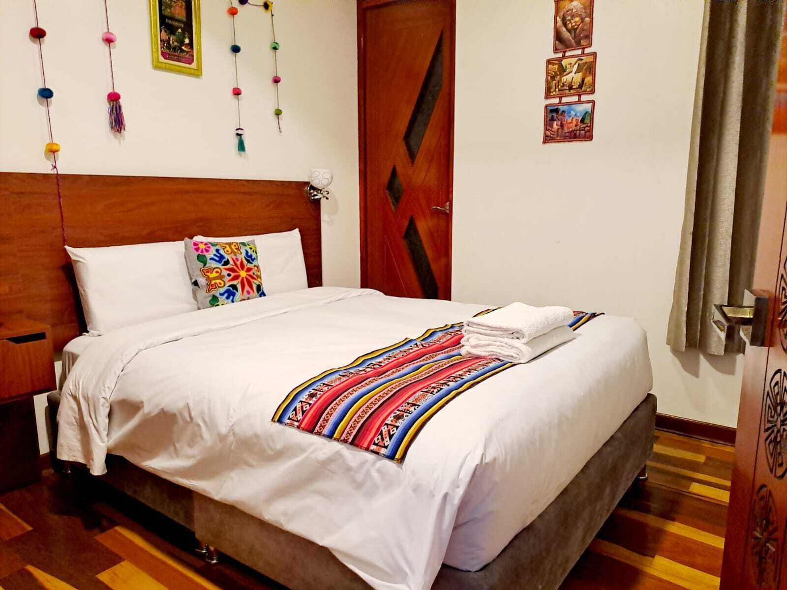 Un letto rifatto con cura, una coperta colorata, una testiera in legno, un piccolo comodino e una porta con un motivo geometrico.