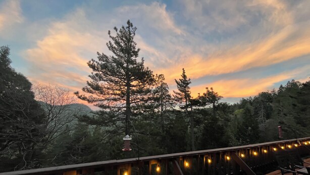 Una terraza de madera con luces colgantes, con vistas a un  área boscosa y un atardecer de fondo.