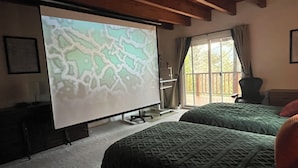 Una habitación con cama, una pantalla proyectora mostrando un mapa, una ventana con vista a árboles y techo de madera.