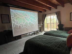 Ein Zimmer mit einem Bett, einer Projektionsleinwand, die eine Karte zeigt, einem Fenster mit Blick auf Bäume und einer Holzbalkendecke.