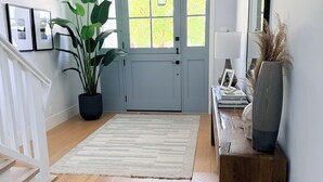 Ein Flur mit Holzboden, einem weißen Teppich, einer Zimmerpflanze, einem Holzkonsolentisch mit einer Vase und einer Tür mit Fenstern.