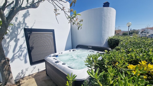 A hot tub in a garden with a white wall, a window with blinds, and a tree.