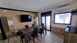 A dining area with a wooden table and chairs, a flat-screen TV mounted on the wall, and a large window with a view of palm trees.