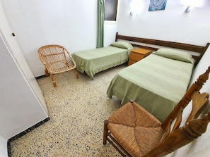 A room with two single beds, a wooden chair, a small wooden table, and a window with a green curtain. - KARINA APARTMENT BEACH-3 (Empuriabrava)