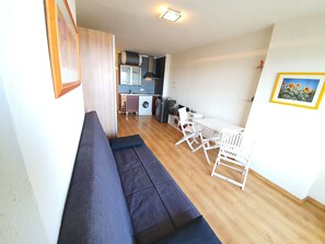 Un espacio habitable compacto con sofá, una mesa de comedor con sillas, una zona de cocina con lavadora y un cuadro en la pared.