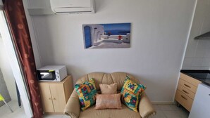 A living room with a beige sofa, colorful cushions, a white air conditioner, a painting of a coastal scene, and a microwave on a wooden cabinet. - APARTMENT YELLOW SEA 173 (Empuriabrava)
