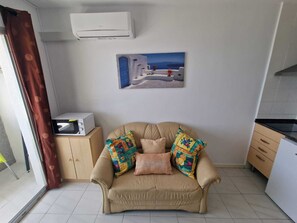 A living room with a beige sofa, colorful cushions, a white air conditioner, a painting of a coastal scene, and a microwave on a wooden cabinet. - APARTMENT YELLOW SEA 173 (Empuriabrava)