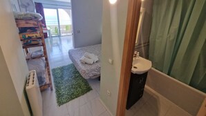 A bedroom with a bed, a green rug, a bedside table, a window, and a bathroom with a sink and shower curtain. - APARTMENT YELLOW SEA 173 (Empuriabrava)