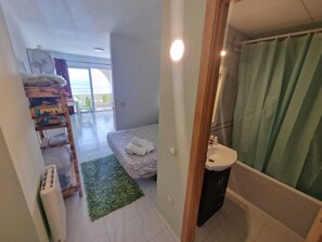 A bedroom with a bed, a green rug, a bedside table, a window, and a bathroom with a sink and shower curtain. - APARTMENT YELLOW SEA 173 (Empuriabrava)