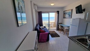 En kompakt opholdsstue med sofa, spisebord, tekøkken og havudsigt gennem en glasdør.