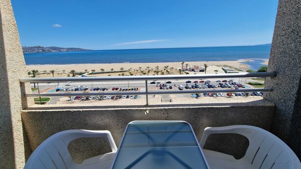 En balkon med et bord og stole, med udsigt over en strand og en parkeringsplads.