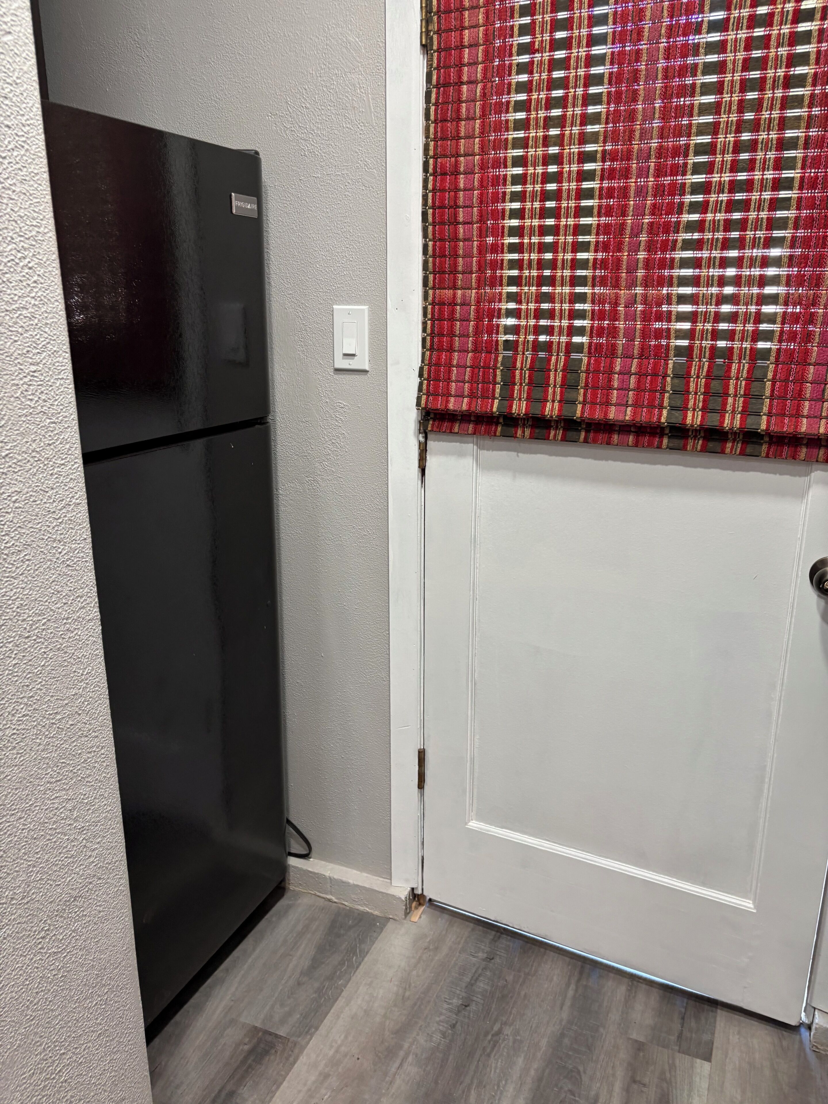 A kitchen with a black refrigerator, a white door, and a red plaid window blind.