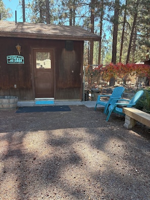 A rustic cabin with a wooden door, a welcome sign, and two blue Adirondack chairs outside.