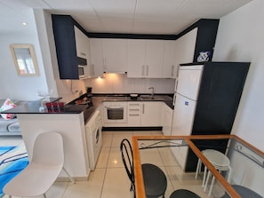 A compact kitchen with white cabinets, a built-in oven, a washing machine, and a refrigerator. - VILLA PROMENADE TERRACE. (Empuriabrava)