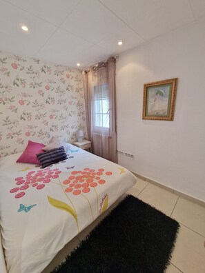 A bedroom with a floral wallpaper, a bed with a floral bedspread, a black rug, a nightstand with a lamp, a window with curtains, and a framed picture on the wall. - VILLA PROMENADE TERRACE. (Empuriabrava)