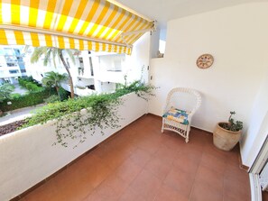 Un balcon agrémenté d’un auvent à rayures, une chaise en osier, un pot de fleurs et une vue sur un immeuble doté de balcons et de verdure.