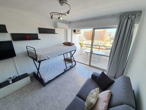 Un espace de vie compact comprenant un lit superposé, un canapé, un téléviseur fixé au mur et un balcon offrant une vue sur les immebles.