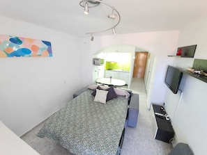 Une petite pièce comprenant un lit, un bureau, une chaise, un téléviseur et, au fond, un coin cuisine.