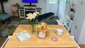 A living room with a TV, a dining table with a book and flowers, a sofa, and a kitchen area.