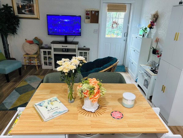 En stue med TV, spisebord med bog og blomster, sofa og et køkkenområde.