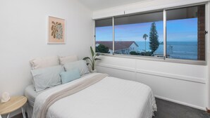 Une chambre avec une grande fenêtre, un lit recouvert de draps blancs, une petite table et un tableau encadré au mur.